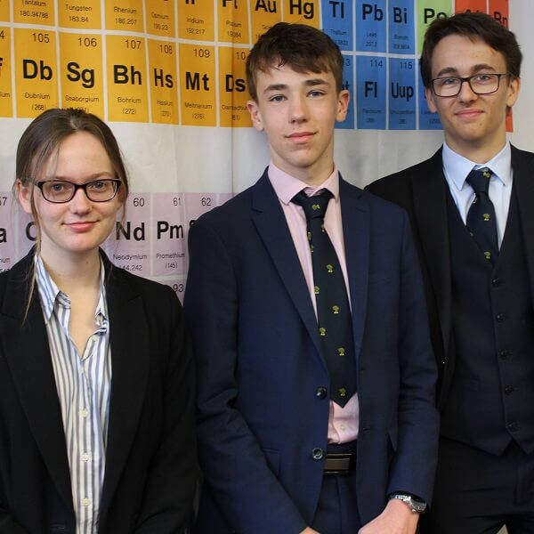 Three Pocklington School students in Sixth Form unifrom stood in front of a poster of the periodic table