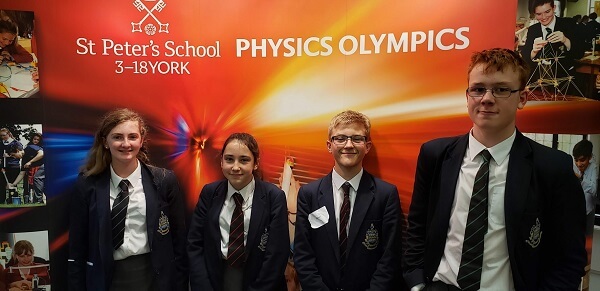 Four Pocklington School pupils stand in front of orange banner showing pictures of pupils engaged in science and the white writing Physics Olympics