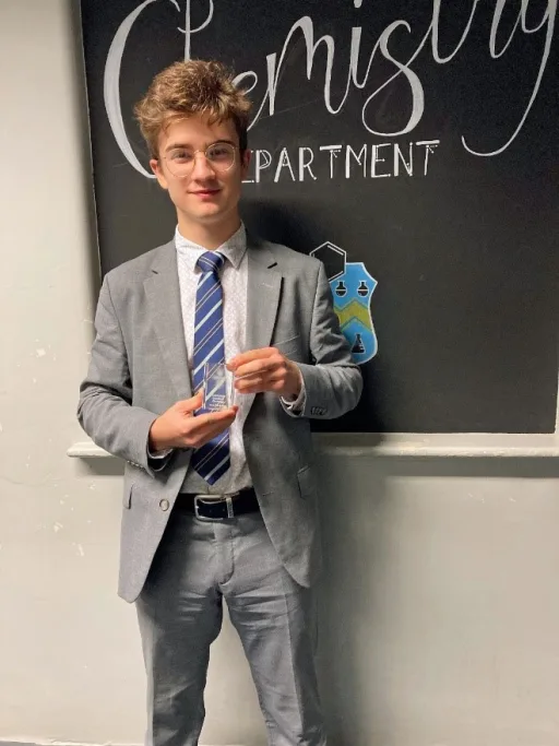 boy stands holding roentgenium chemistry award infront of chemistry department sign