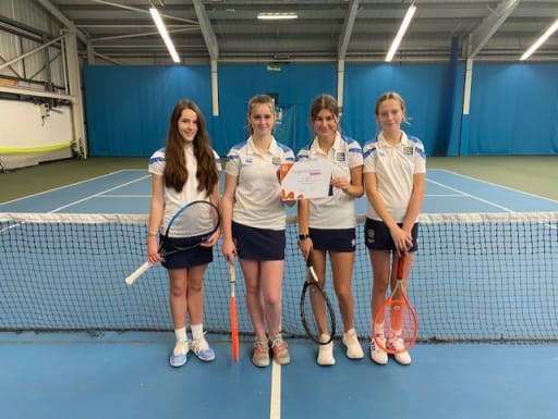 U13 girls tennis squad stood on an indoor tennis court holding rackets and competition certificate